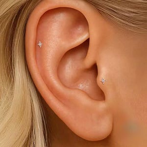 May include: Two small, silver-toned star-shaped stud earrings are positioned in the ear. One earring is located on the upper cartilage, and the other is placed on the tragus. Each stud features a four-pointed star design with a tiny central stone.