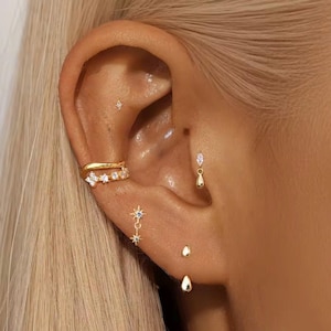 May include: A collection of gold-toned earrings including a star-shaped stud, a double-band ear cuff with clear stones, a marquise-cut stone stud, a dangling star charm, and two smooth teardrop-shaped studs worn in various ear piercings.