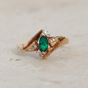 May include: A gold ring with a marquise-cut emerald center stone and small diamond accents. The ring has a unique, angular design. The emerald is a vibrant green, and the diamonds sparkle. A piece of jewelry.