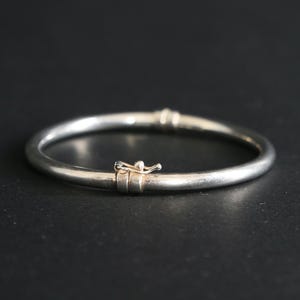 Może przedstawiać: Srebrna bransoletka typu obręcz ze zapięciem. Okrągła bransoletka ma gładką, wypolerowaną powierzchnię. Zapięcie znajduje się z przodu i wydaje się być wykonane z metalu. Średnica wynosi około 6,5 cm.