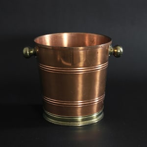 May include: A copper-coloured bucket with two small brass handles. The bucket has a brass base and two horizontal bands around the body. The interior of the bucket is reflective, set against a black background.