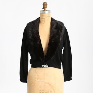 May include: A black cropped cardigan with a faux fur collar. The cardigan has a V-neck and a single button closure. The mannequin is wearing the cardigan on a white background.