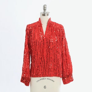 Può includere: Top rosso brillante con paillettes, maniche lunghe e scollo a V. L'indumento è esposto su un manichino con la scritta "COLLAPSIBLE SHOULDER" e il numero "6". Il top è un capo d'abbigliamento accattivante.