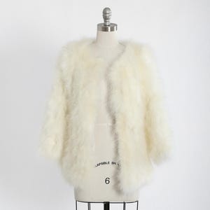 Könnte beinhalten: Elfenbeinfarbene, federartige Jacke auf einer Schaufensterpuppe. Die Jacke hat einen runden Ausschnitt und Dreiviertelärmel. Die Schaufensterpuppe ist Größe 6 und wird von einem schwarzen Metallständer getragen. Der Hintergrund ist schlicht weiß.