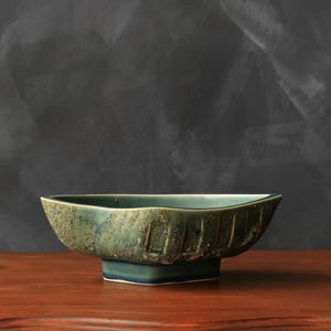 May include: A ceramic bowl with a teal and green glaze featuring a textured, speckled exterior surface and vertical grooved detailing. The bowl has a wide, open top with a slightly irregular rim and sits on a square, raised base.
