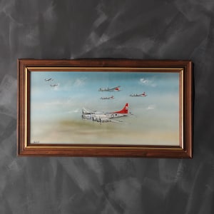 Op de afbeelding: Een ingelijst schilderij van verschillende vintage vliegtuigen in de lucht tegen een blauwe lucht met wolken. Het centrale vliegtuig is zilverkleurig met een rode staart. De lijst is donker hout.
