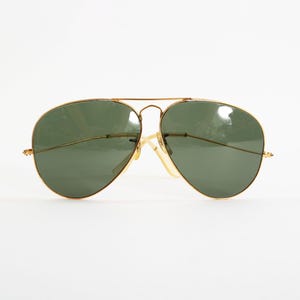 May include: Aviator sunglasses featuring a thin gold-tone metal frame and dark green teardrop-shaped lenses. The design includes a double bridge across the top and slender temples with clear nose pads, providing a classic look with a metallic finish.