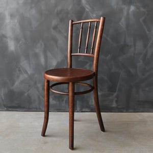 May include: A dark brown wooden chair featuring a circular seat, a curved backrest with three vertical spindles, and four slightly flared legs connected by a circular support ring beneath the seat, showcasing a smooth, polished wood grain finish.