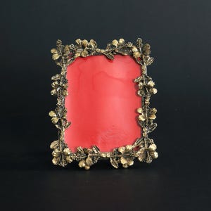 May include: A rectangular antique brass picture frame with a red interior. The frame is adorned with floral and leaf detailing around the edges. The frame is set against a dark background.
