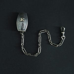 May include: A silver pocket watch chain with a rectangular fob featuring the letter "E". The chain is composed of elongated links and terminates in a clasp. The fob and chain are set against a black background.