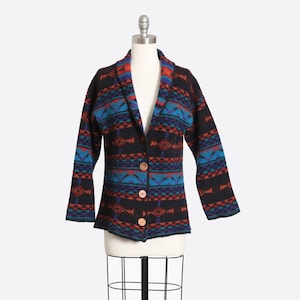 May include: A long-sleeve cardigan featuring a shawl collar and three light brown buttons. The knit fabric displays a repeating geometric pattern in shades of black, bright blue, and burnt orange across the entire garment.