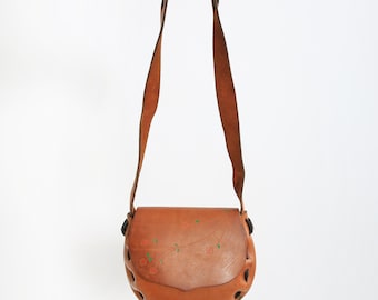 Peut inclure: Sac bandoulière en cuir marron clair avec un motif floral. Le sac a une forme arrondie, une longue bandoulière et des découpes décoratives sur le bord inférieur. Le motif floral présente des accents rouges et verts.