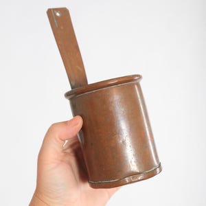 May include: A copper measuring cup with a long, flat handle. The cup is cylindrical with a slightly flared rim and a riveted base. The handle has a hole near the top. The copper has a warm, reddish-brown hue.