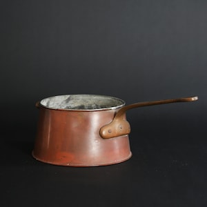 May include: A copper saucepan with a long handle. The saucepan has a slightly flared lip and a worn, aged patina.