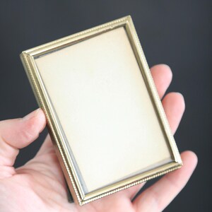 May include: A small, rectangular gold-colored picture frame with a beaded edge. The frame is held in a hand against a dark background. The frame is empty, with a light-colored interior.