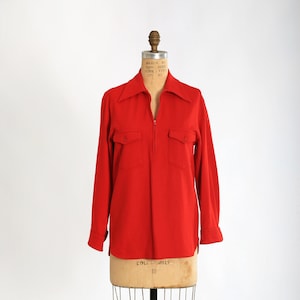 Puede incluir: Una camisa roja de manga larga con cierre de cremallera y dos bolsillos de solapa. La camisa tiene un cuello ancho y se muestra en un maniquí.