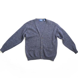 Può includere: Un cardigan blu con scollo a V e chiusura con bottoni. Il maglione ha maniche lunghe e due tasche anteriori. Il tessuto ha una trama maculata. L'indumento è steso su una superficie bianca.