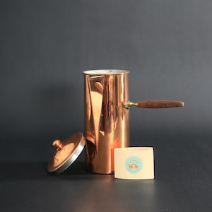 May include: A copper coffee pot with a wooden handle and a lid. The pot is cylindrical and has a shiny finish. A small rectangular card with a logo is next to the pot. The background is a dark gray.
