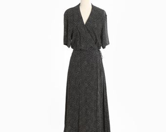 May include: A black wrap dress with a white polka dot pattern. The dress has short sleeves and a V-neckline. It is displayed on a mannequin against a white background. The dress is floor-length and has a tie closure at the waist.