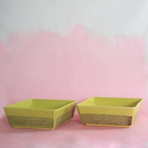 Peut inclure: Deux bols carrés en céramique avec un glaçage jaune et une bande dorée autour du haut. Les bols sont posés sur une surface rose.