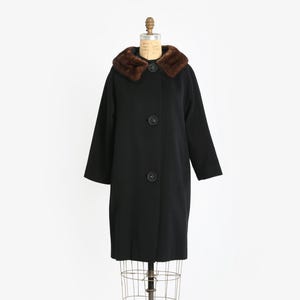 May include: A vintage black coat with a brown fur collar and three large black buttons. The coat is displayed on a dress form against a white background. The coat has long sleeves and a classic silhouette.