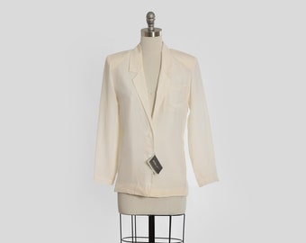 Puede incluir: Una chaqueta blanca de lino con un cierre de botón único y un bolsillo en el lado izquierdo. La chaqueta está en un maniquí blanco.