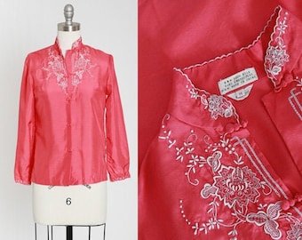 May include: A pink silk blouse with white embroidery featuring floral designs. The blouse has a mandarin collar and long sleeves. The label reads "100% Silk, Dry Clean Only, Made in China".