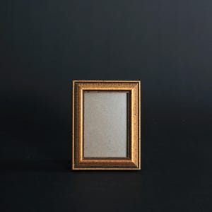 May include: A small, rectangular picture frame with a gold-colored, ornate border. The frame has a neutral-colored interior, and is set against a dark background. The frame is likely for displaying a photograph or artwork.