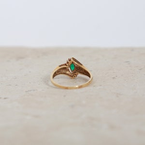 May include: A gold ring with a marquise-cut green gemstone at the center. The ring has a V-shaped design with small decorative elements on either side of the gemstone. The ring is set against a neutral background.