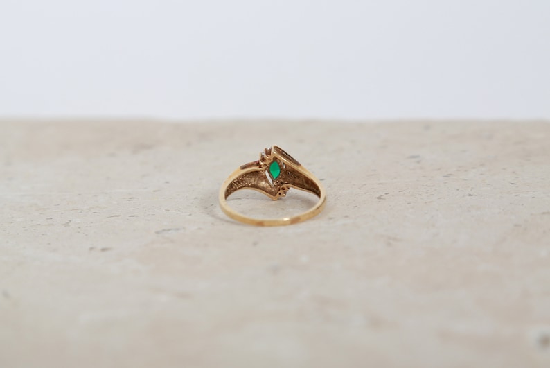 May include: A gold ring with a marquise-cut green gemstone at the center. The ring has a V-shaped design with small decorative elements on either side of the gemstone. The ring is set against a neutral background.