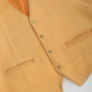May include: A yellow leather vest with a pointed collar and multiple button closures. The vest has two pockets on the front and a smooth, textured surface.