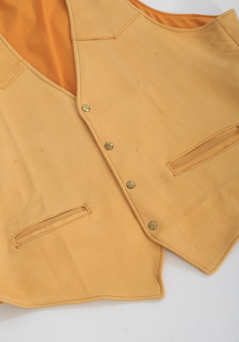 May include: A yellow leather vest with a pointed collar and multiple button closures. The vest has two pockets on the front and a smooth, textured surface.
