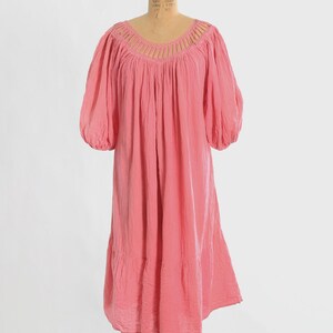 K&ouml;nnte beinhalten: Ein knielanges rosa Kleid mit einem weiten, abgerundeten Ausschnitt und vertikalen Ausschnittdetails. Das Kleidungsst&uuml;ck hat volumin&ouml;se, ellbogenlange Puff&auml;rmel und einen gerafften, gestuften Rock, der eine lockere Silhouette erzeugt.