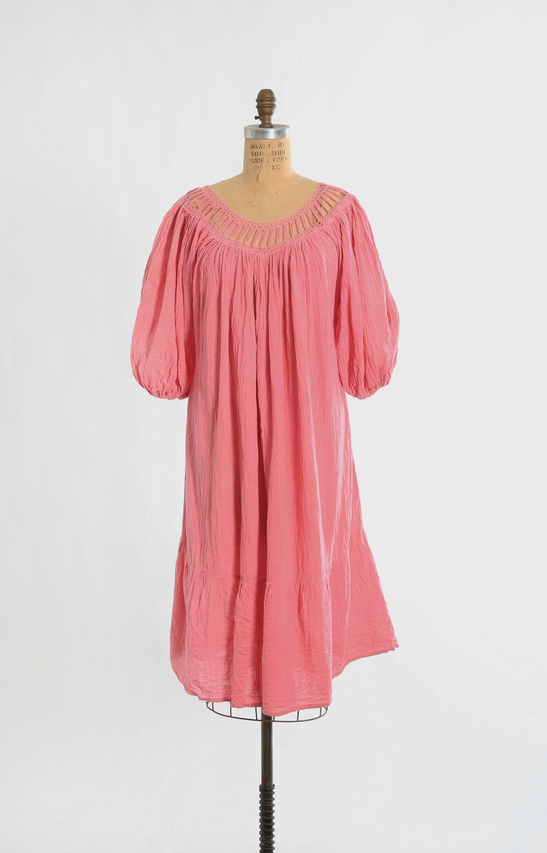 K&ouml;nnte beinhalten: Ein knielanges rosa Kleid mit einem weiten, abgerundeten Ausschnitt und vertikalen Ausschnittdetails. Das Kleidungsst&uuml;ck hat volumin&ouml;se, ellbogenlange Puff&auml;rmel und einen gerafften, gestuften Rock, der eine lockere Silhouette erzeugt.