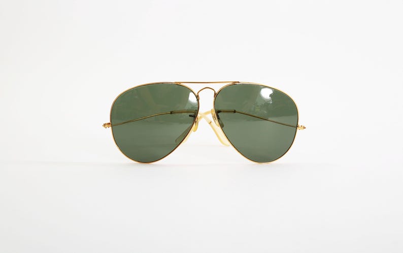 May include: Aviator sunglasses featuring a thin gold-tone metal frame and dark green teardrop-shaped lenses. The design includes a double bridge across the top and slender temples with clear nose pads, providing a classic look with a metallic finish.