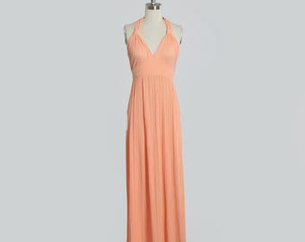 May include: A peach-colored, floor-length halter dress on a mannequin. The dress features a V-neckline and a pleated skirt. The dress is displayed on a black rolling stand against a light gray background.