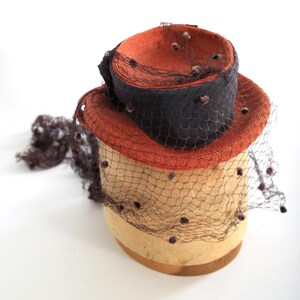 May include: A vintage hat with a brown and orange straw base and a black ribbon. The hat is on a hat form and is covered in a black net with brown dots.