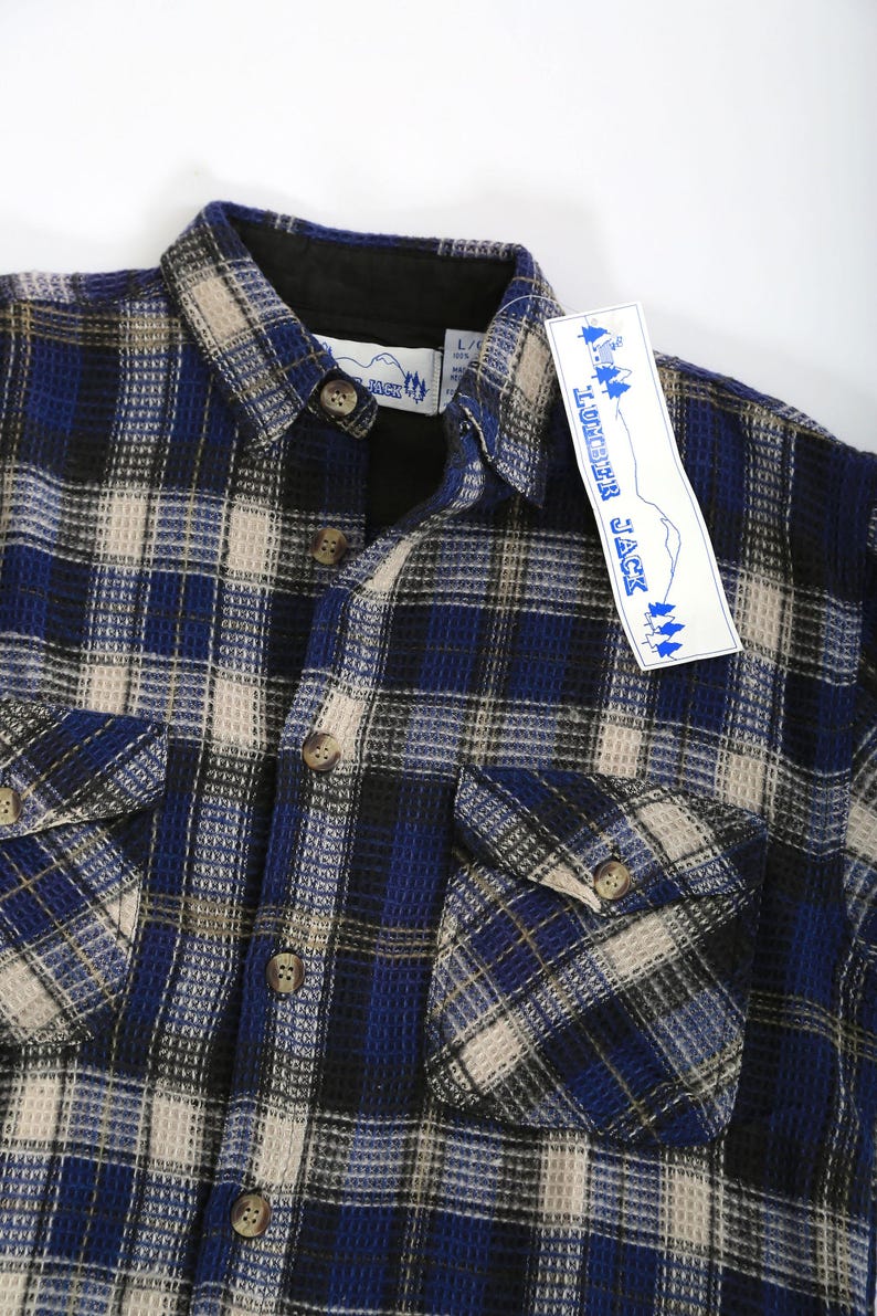 May include: A blue, black, and beige plaid flannel shirt with a woven texture. The shirt has a button-down collar and two chest pockets. The shirt has a "Lumber Jack" tag attached to it.