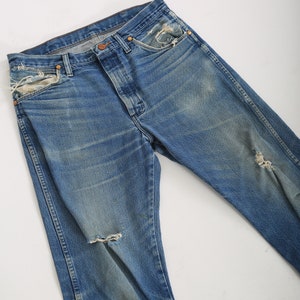 Op de afbeelding: Een paar blauwe denim jeans met distressed details, waaronder scheuren en rafelige randen. De jeans heeft een klassieke rechte pijp.