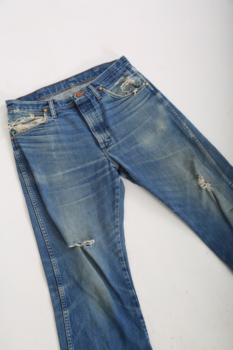 Op de afbeelding: Een paar blauwe denim jeans met distressed details, waaronder scheuren en rafelige randen. De jeans heeft een klassieke rechte pijp.