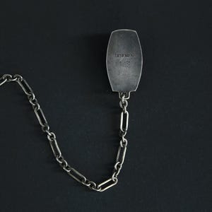 May include: A silver-toned metal tie clip with a chain. The rectangular clip has rounded edges and is connected to a chain of elongated links. The clip is stamped with the words "HITOROK" and "SILVER FRONT".