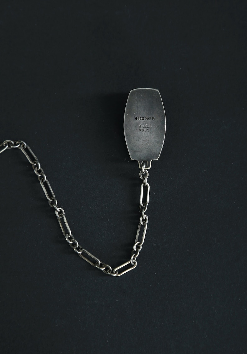 May include: A silver-toned metal tie clip with a chain. The rectangular clip has rounded edges and is connected to a chain of elongated links. The clip is stamped with the words "HITOROK" and "SILVER FRONT".