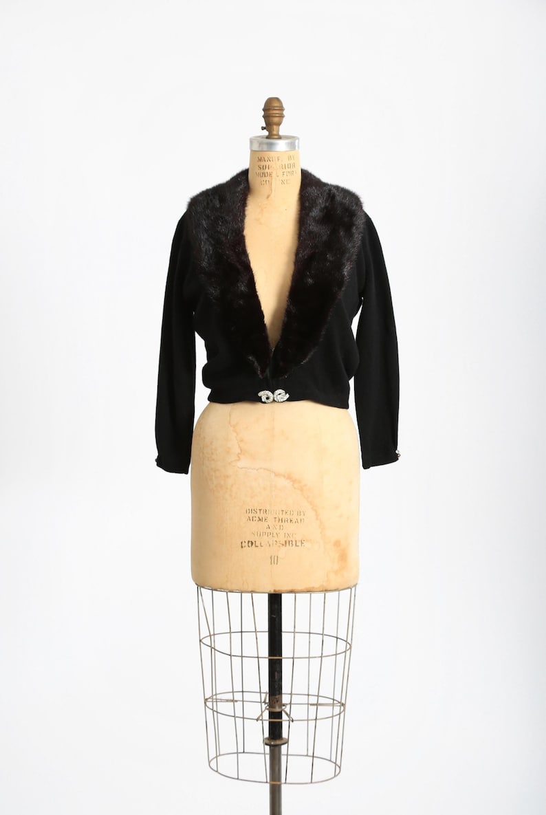 May include: A black cropped cardigan with a faux fur collar. The cardigan has a V-neck and a single button closure. The mannequin is wearing the cardigan on a white background.