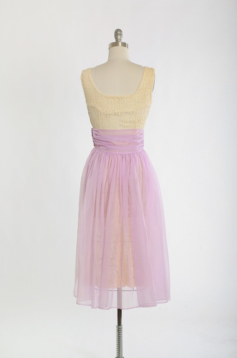Op de afbeelding: Een vintage jurk zonder mouwen met een geel kanten lijfje en een lavendel tule rok. De jurk heeft een getailleerd lijfje en een volle rok.