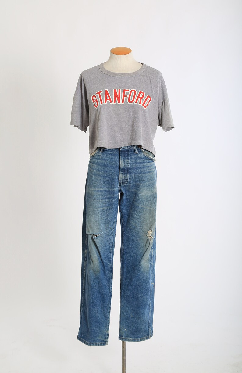 Op de afbeelding: Een grijs gecropped t-shirt met het woord "STANFORD" in rood gedrukt op de voorkant, gecombineerd met een blauwe denim jeans met distressed details.