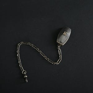 May include: A silver-toned pocket watch fob with a rectangular case and a chain. The case has a clear, possibly glass, face with a small design. The chain is made of small, linked pieces.