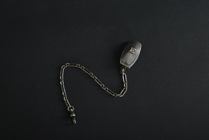 May include: A silver-toned pocket watch fob with a rectangular case and a chain. The case has a clear, possibly glass, face with a small design. The chain is made of small, linked pieces.