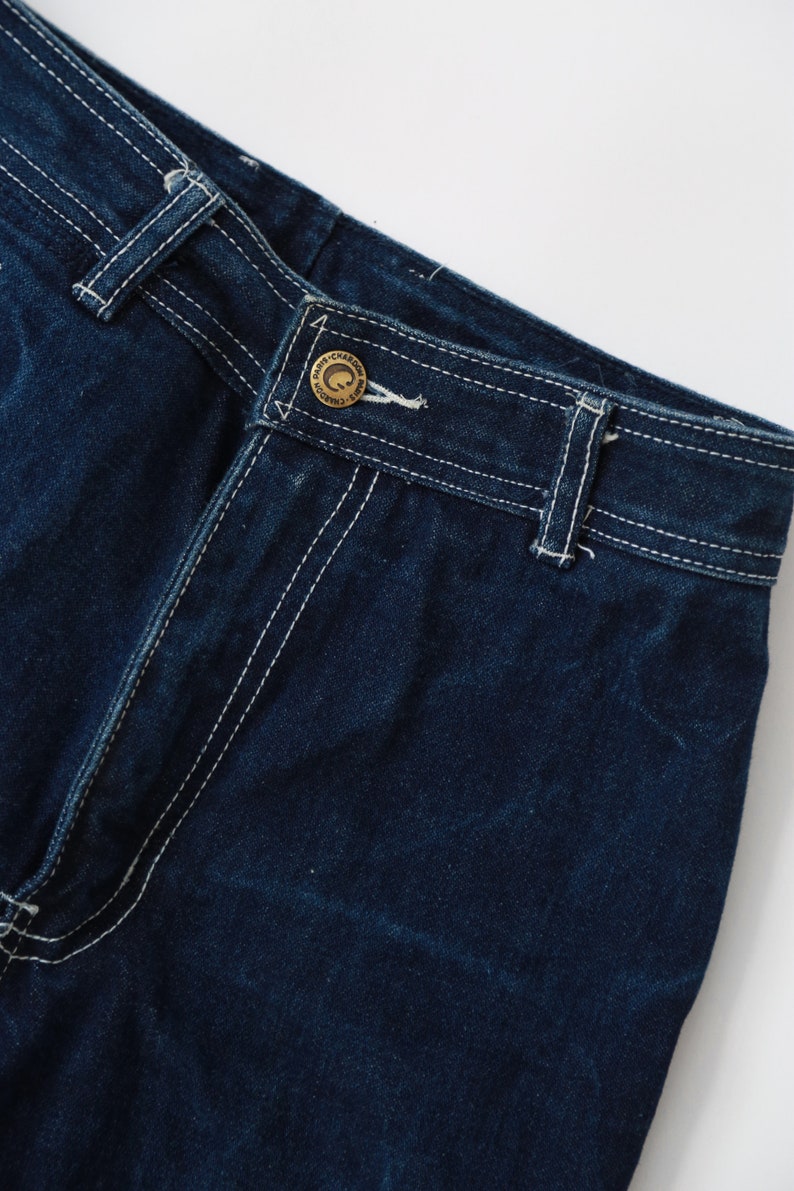 May include: Close-up of dark blue denim jeans with white stitching and a gold button with the text "CHARCOAL" on it.