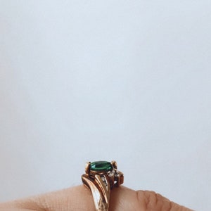 May include: Gold-toned ring featuring a large, oval-cut emerald green gemstone as the centerpiece. The ring has a detailed, ornate design with smaller accent stones. The ring is set on a finger.