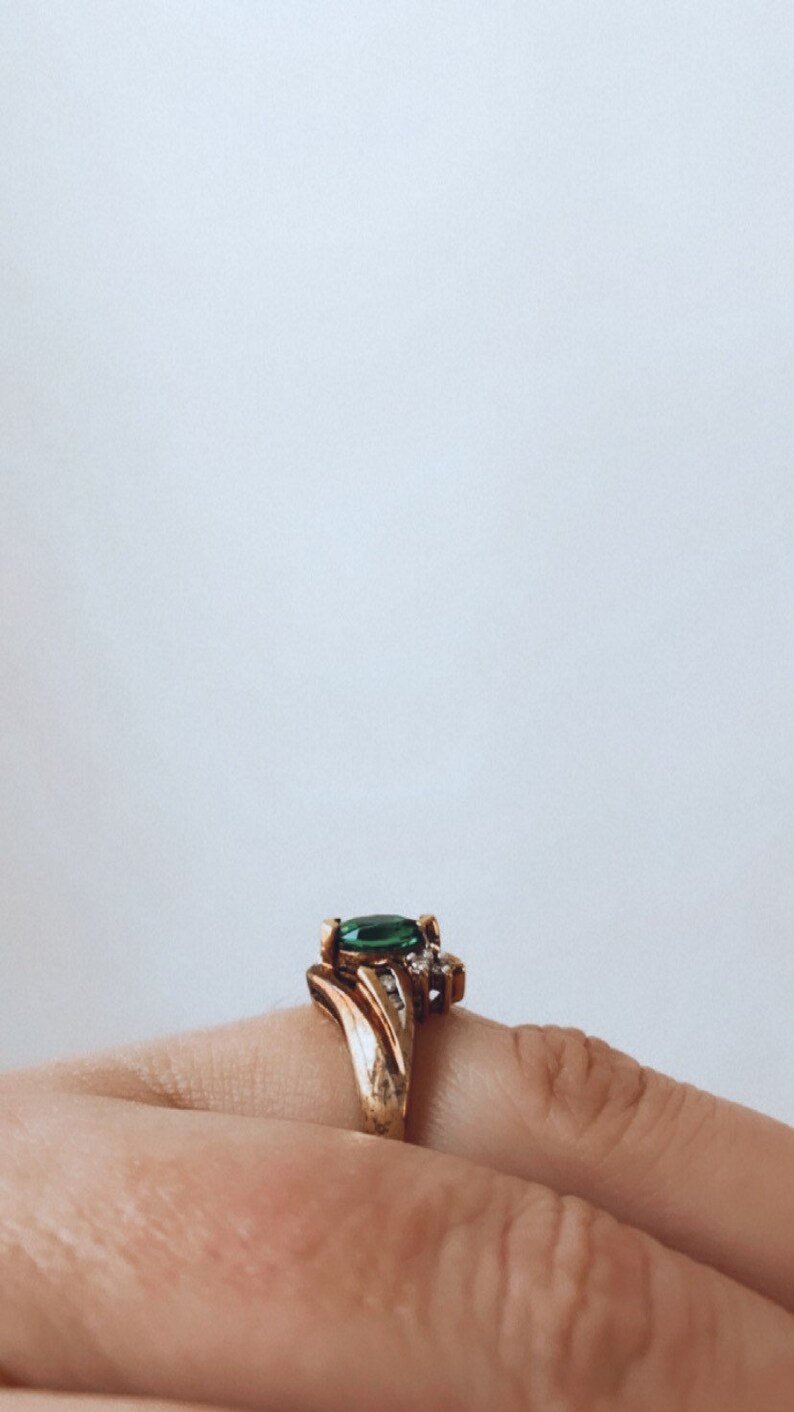 May include: Gold-toned ring featuring a large, oval-cut emerald green gemstone as the centerpiece. The ring has a detailed, ornate design with smaller accent stones. The ring is set on a finger.